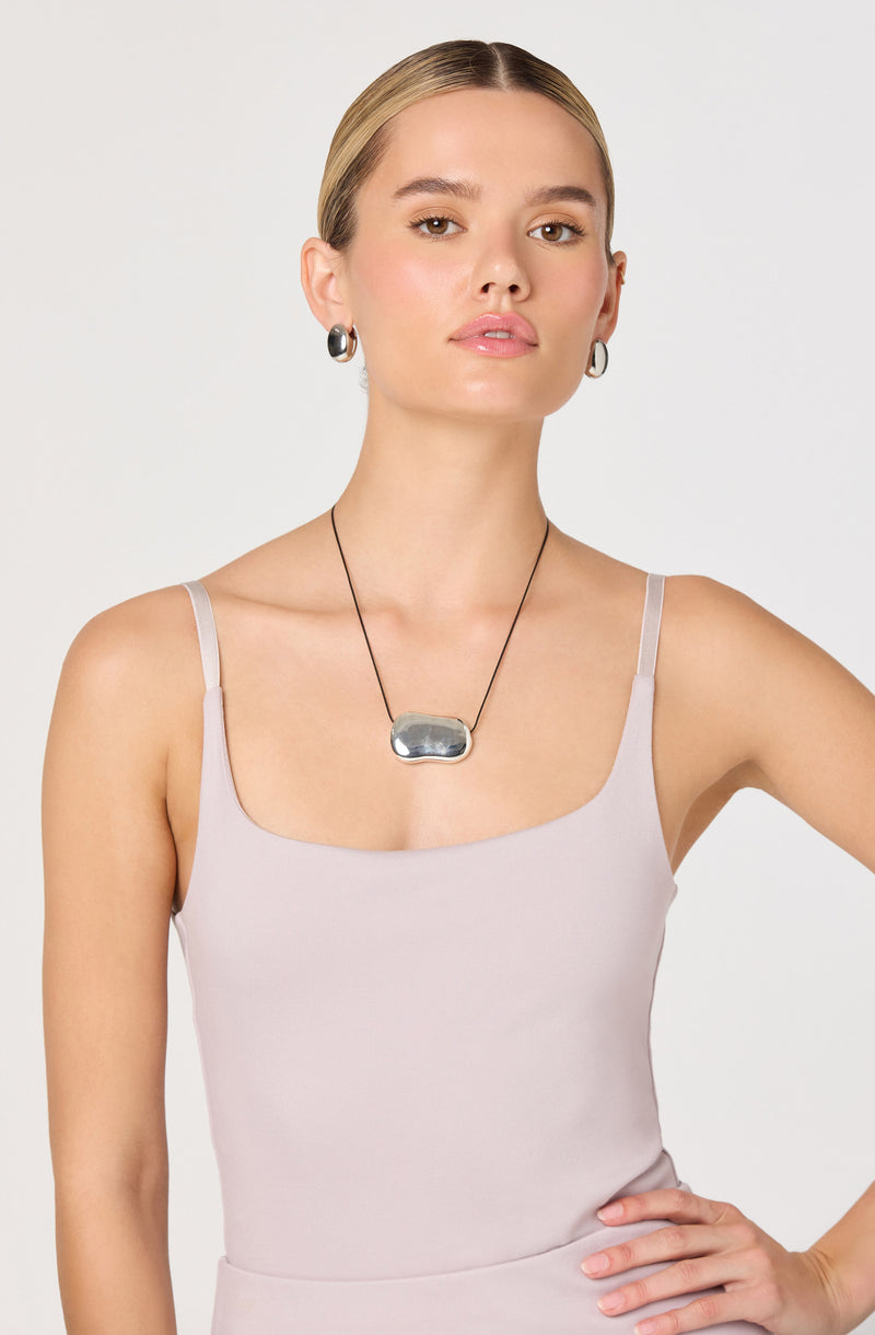 The woman is wearing a light-colored, sleeveless camisole with thin spaghetti straps. She has a silver, rounded rectangular pendant necklace on a black cord, and is accessorized with matching silver hoop earrings.