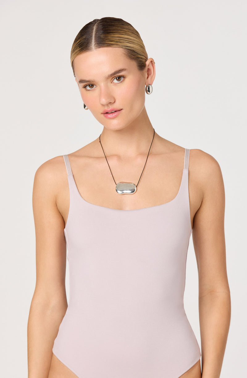 A woman is wearing a light beige or cream-colored sleeveless bodysuit with thin spaghetti straps. She has accessorized with large hoop earrings and a chunky silver pendant necklace on a black cord. Her hair is styled in a sleek, middle-parted bun, and she has a neutral, slightly smiling expression. The background is plain and light-colored.
