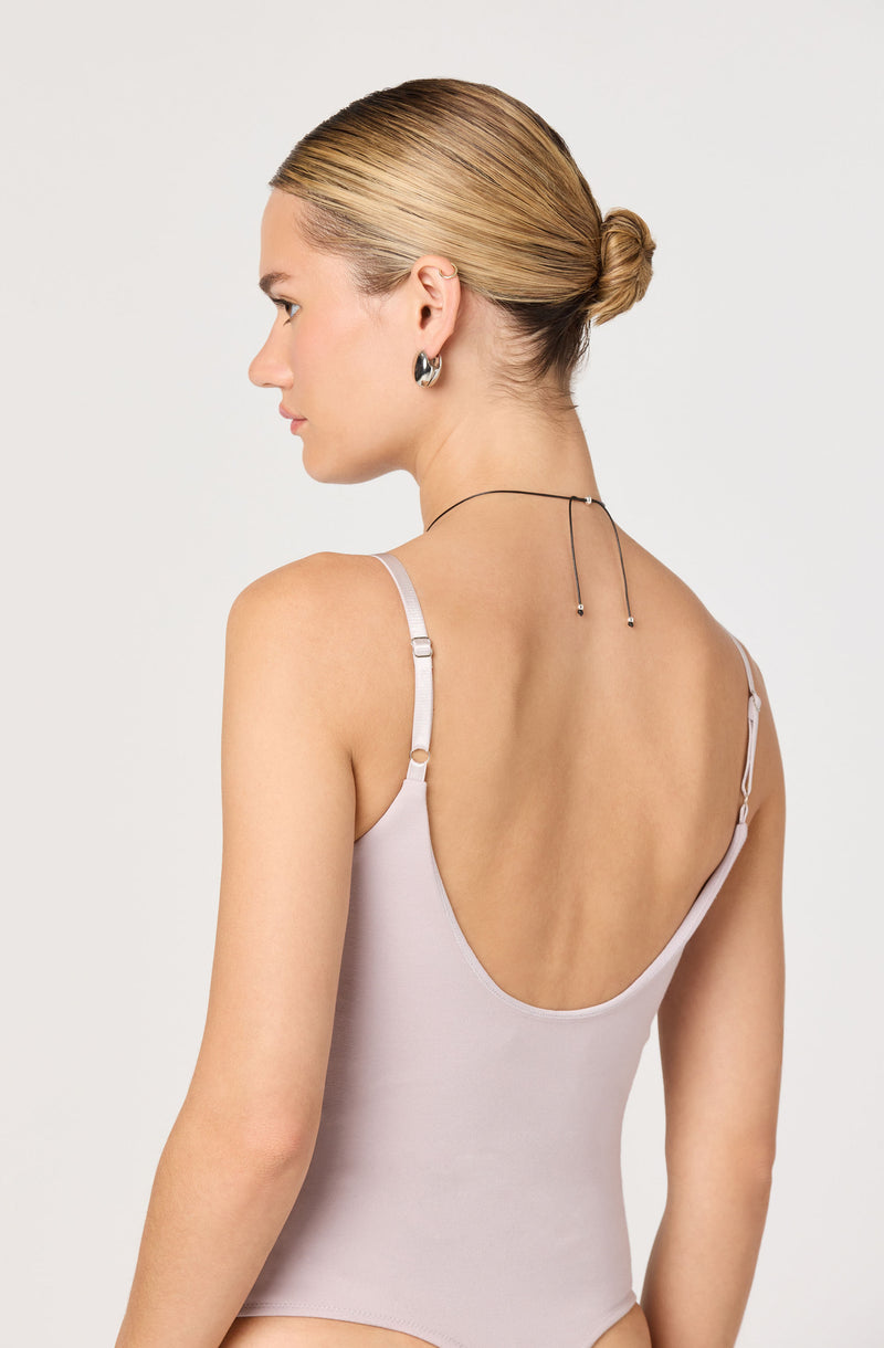 A woman is wearing a light pink sleeveless camisole with adjustable shoulder straps. She has blonde hair styled in a low bun and is accessorized with hoop earrings and a delicate black necklace with small beads.