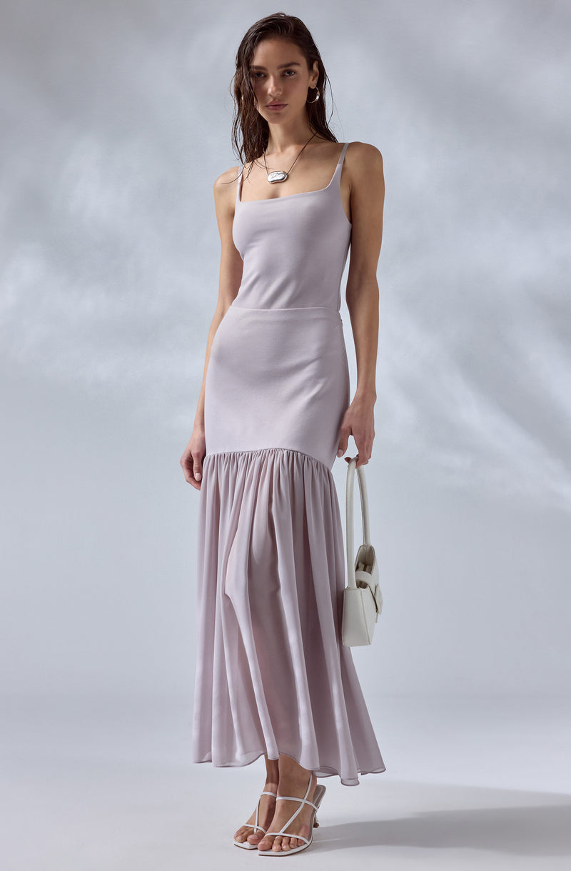 A woman is wearing a light lavender, sleeveless dress with spaghetti straps. The dress has a fitted bodice and transitions into a tiered skirt, with the lower part made of a flowy, sheer fabric gathered at the seam. She is accessorizing with hoop earrings, a necklace, and white strappy high-heeled sandals. She is carrying a small, white handbag.