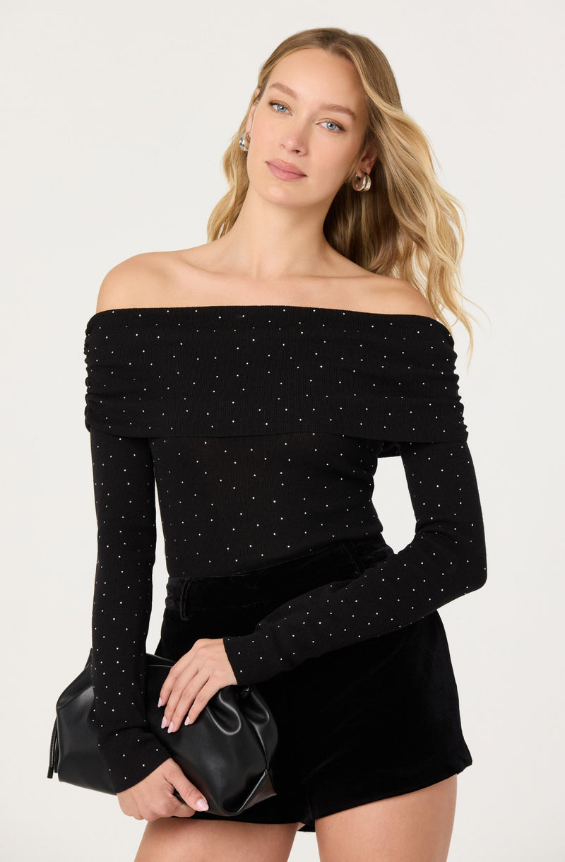 A woman is wearing an off-the-shoulder black top with long sleeves, decorated with small white polka dots. She is also wearing black high-waisted shorts and holding a black clutch purse. She has blonde wavy hair and is accessorized with hoop earrings.