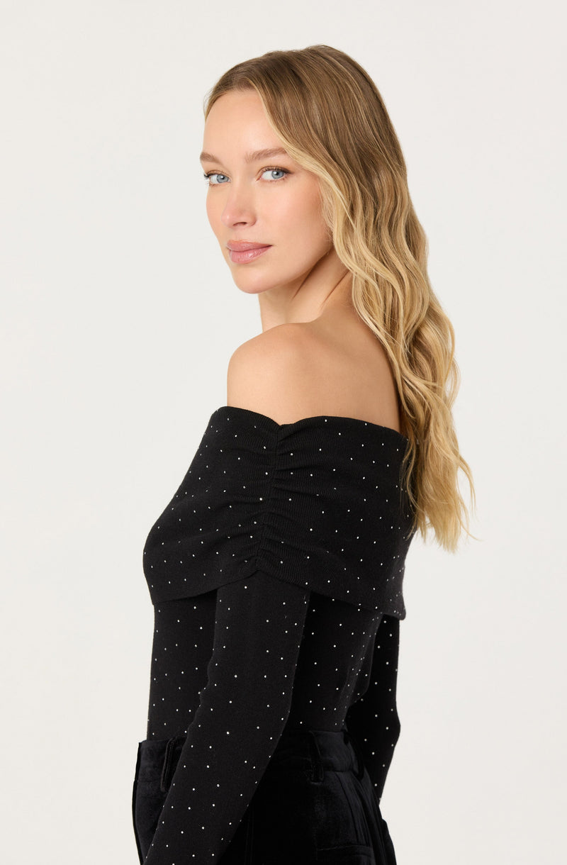 A woman is wearing an off-the-shoulder black top with long sleeves. The top is decorated with small white polka dots all over.
