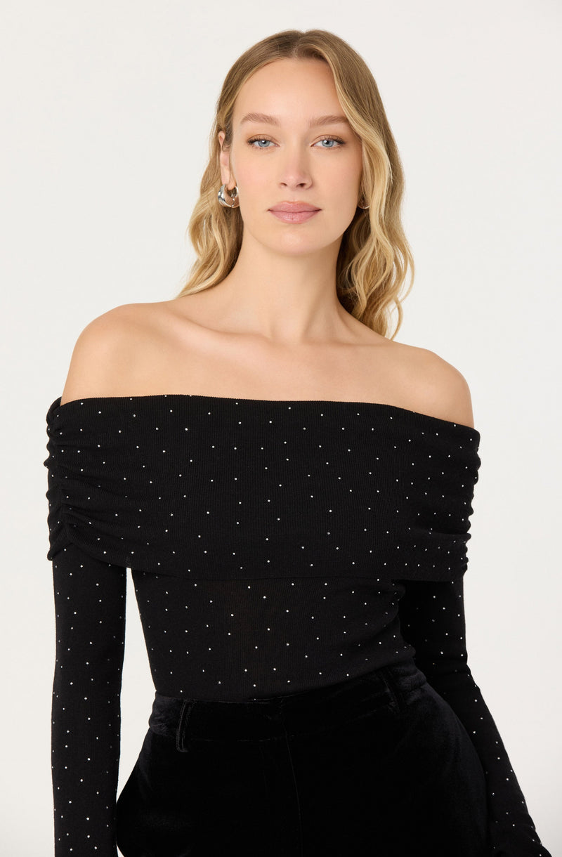 A woman is wearing an off-the-shoulder black top adorned with small white polka dots. The top features a ruched design along the shoulders and sleeves, creating a gathered effect.