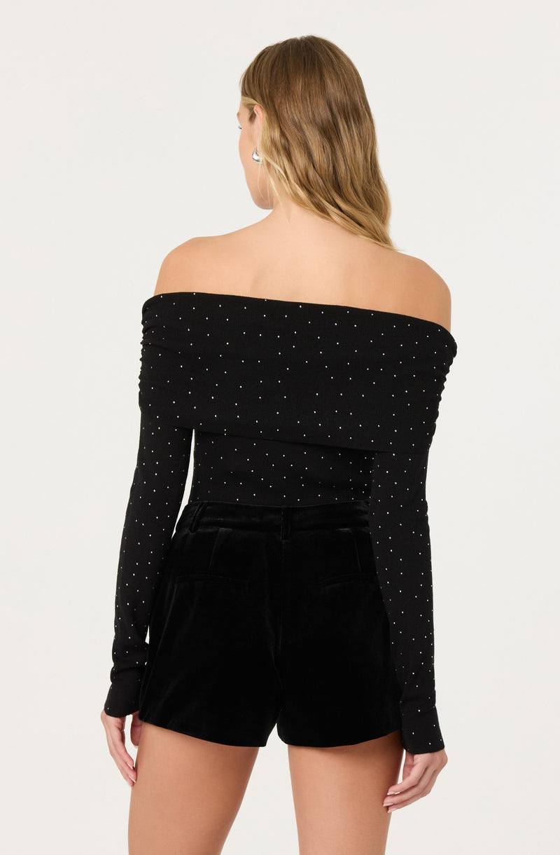 A woman wearing a black off-the-shoulder long-sleeve top with small white polka dots, paired with high-waisted black shorts.