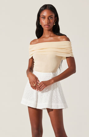 A woman is wearing an off-the-shoulder, cream-colored top with gathered detailing across the chest and sleeves. She pairs it with high-waisted white shorts that have a subtle, eyelet pattern.