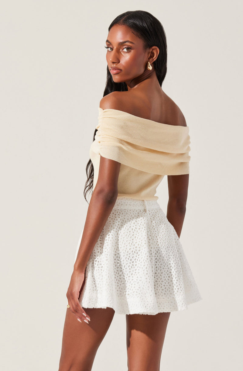 A woman is wearing an off-the-shoulder, cream-colored top with soft, draped fabric. She pairs it with a white, high-waisted skirt featuring a textured, eyelet pattern. She accessorizes with gold hoop earrings and has long, dark hair styled in loose waves.