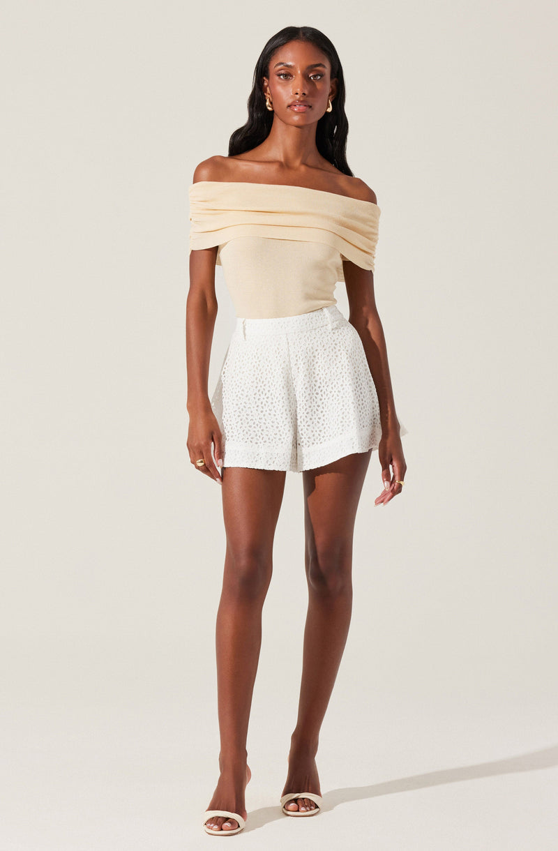 A woman is wearing an off-the-shoulder, cream-colored top with draped, layered fabric around the neckline. She pairs it with high-waisted white shorts that have a subtle textured or dotted pattern. She completes her outfit with white open-toe heels.