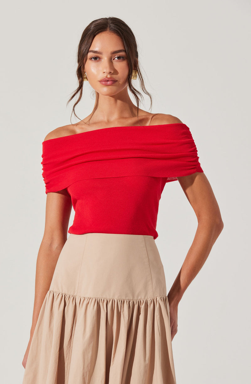 A woman is wearing an off-the-shoulder, bright red top with a textured, ruched design across the chest. She pairs it with a beige, high-waisted skirt that features a gathered, ruffled hem.