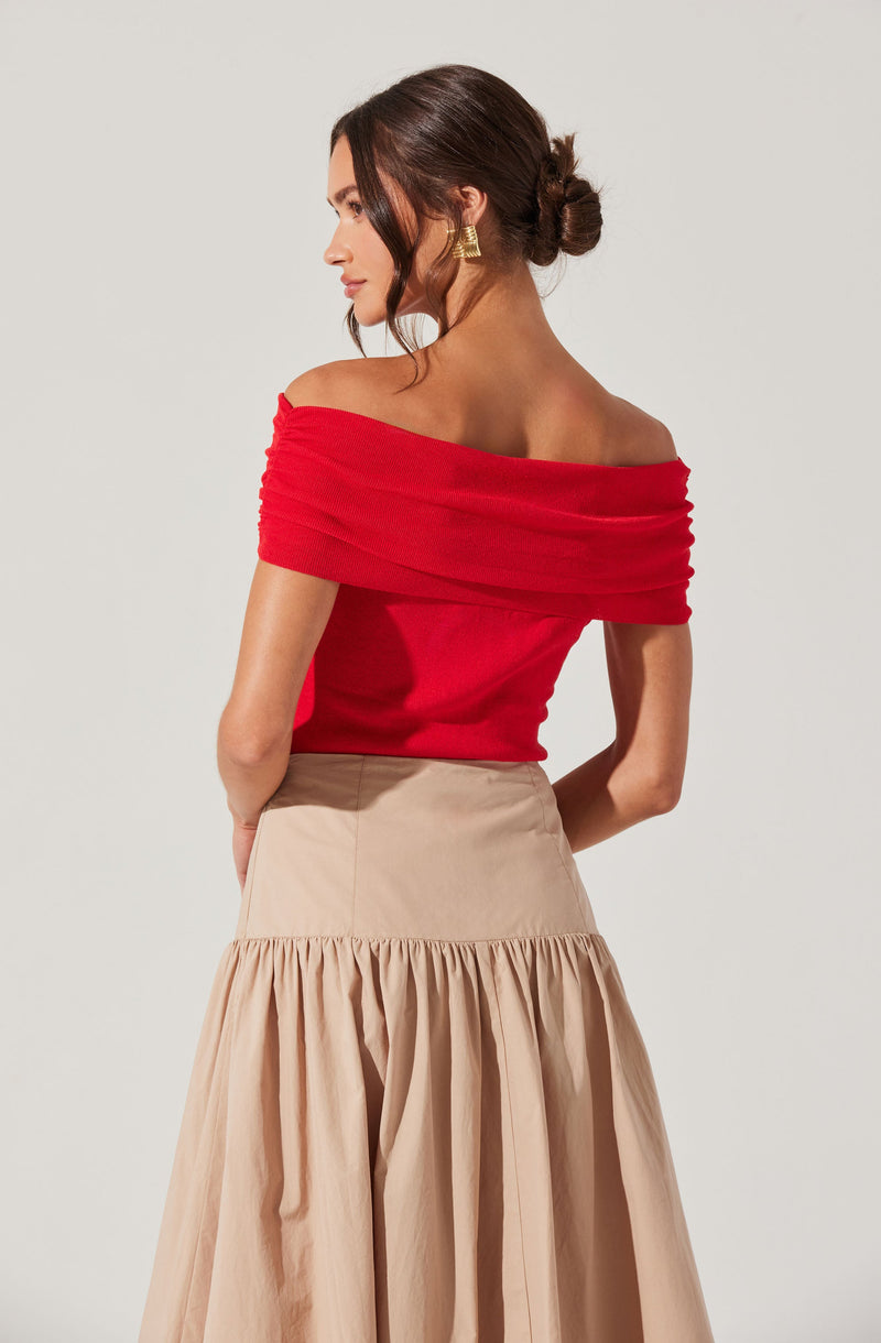 A woman is wearing an off-the-shoulder red top with ruched fabric at the collar, paired with a high-waisted, beige, gathered skirt that has a full, pleated design. She has her hair styled in a loose bun and is accessorized with gold earrings.