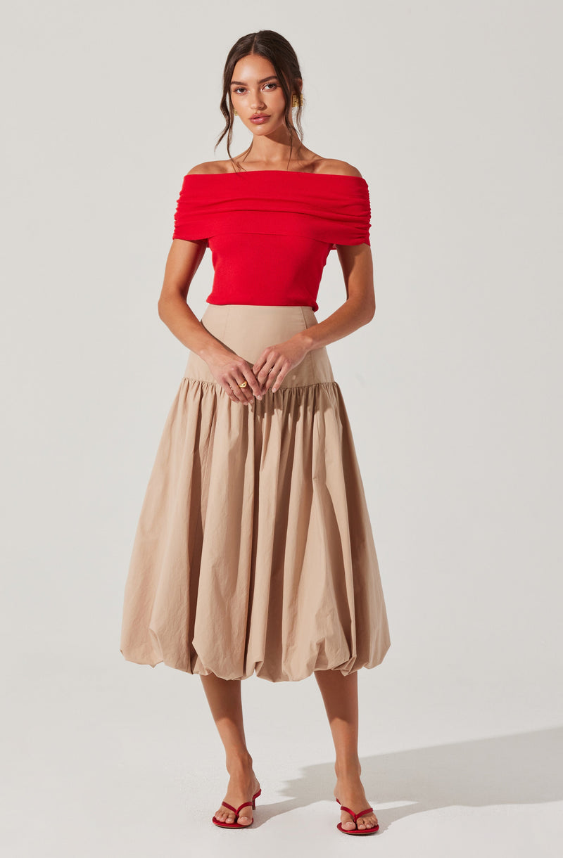 A woman is wearing an off-the-shoulder, bright red top with a fitted bodice and horizontal ruched detailing across the chest and sleeves. She pairs it with a high-waisted, beige, bubble-style midi skirt that has a gathered, voluminous silhouette. She is also wearing red strappy high-heeled sandals.