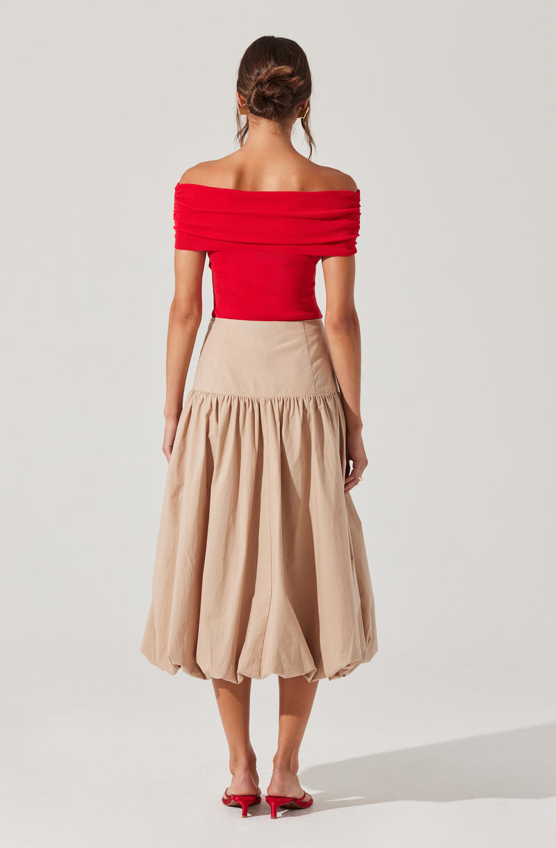 A woman is wearing an off-the-shoulder red top with gathered fabric along the neckline and sleeves, paired with a high-waisted, beige, gathered midi skirt. She is also wearing red high-heeled shoes.