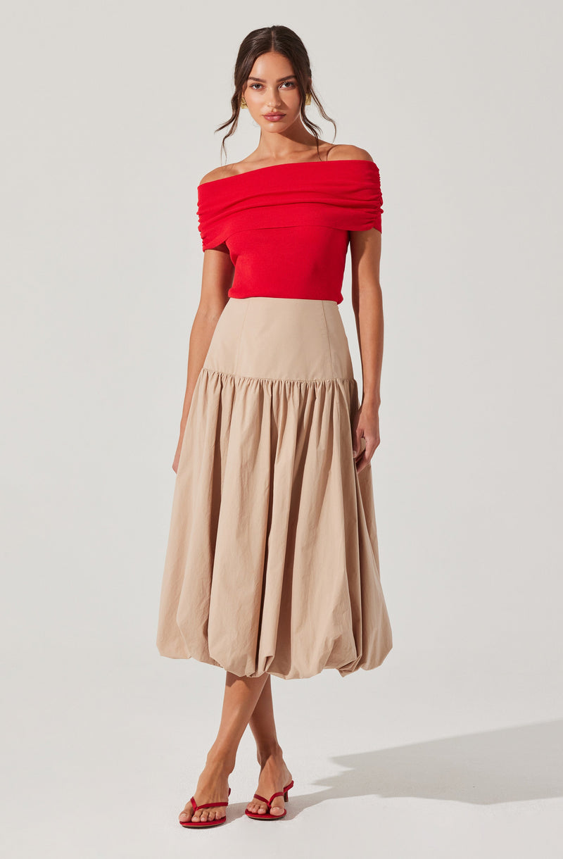 A woman is wearing a bright red, off-the-shoulder top with ruched detailing along the neckline and sleeves. She pairs it with a beige, high-waisted, full-length skirt that has a gathered, voluminous design. She is also wearing red heeled sandals.