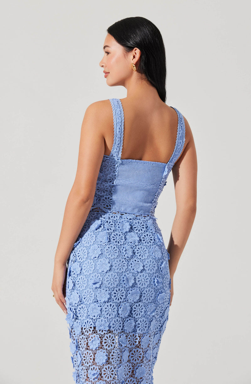 A woman is wearing a sleeveless, blue lace dress with intricate floral patterning. The dress features wide straps and a fitted bodice, with the lace extending from the waist down to the hem, showcasing floral cutouts throughout. She has dark, straight hair and is wearing gold hoop earrings.