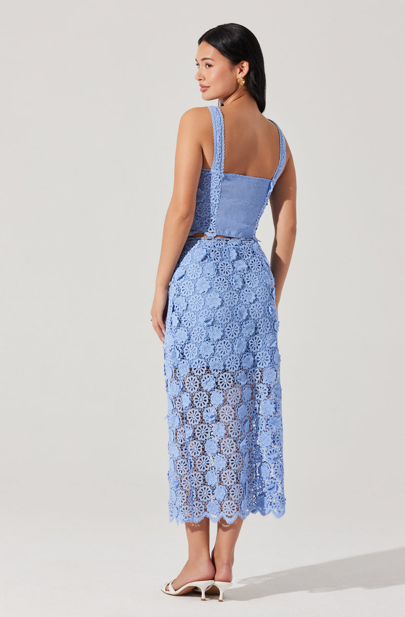 A woman is wearing a two-piece outfit made of blue lace. The top is a sleeveless, fitted tank with wide straps and a square neckline. The matching long skirt is also made of blue lace with a floral pattern, fitted at the waist and flowing down to her ankles with a scalloped hem. She is wearing white open-toe heels.