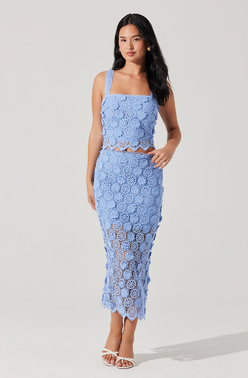 A woman is wearing a two-piece light blue crochet outfit consisting of a sleeveless crop top with wide shoulder straps and a matching high-waisted skirt that reaches mid-calf. The crochet pattern features floral motifs and openwork details. She is also wearing white strappy high-heeled sandals.