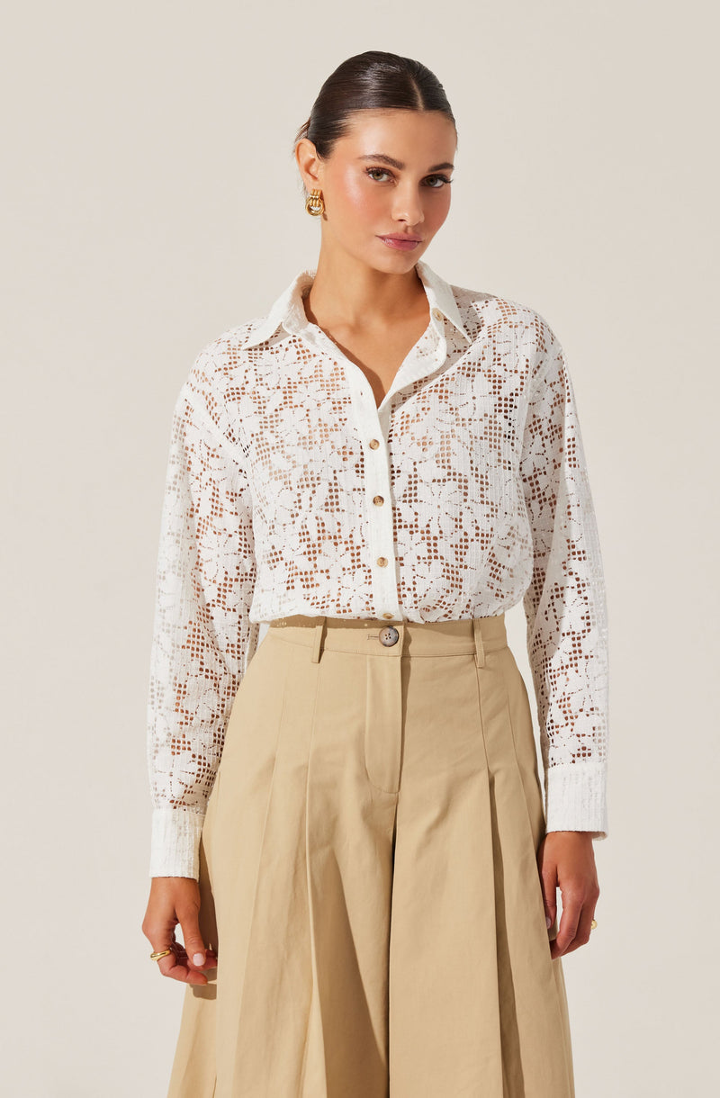 A woman is wearing a white, long-sleeved button-up shirt made of lace with a floral pattern. The shirt has a standard collar and white buttons down the front. She pairs it with high-waisted, wide-leg beige trousers that feature pleats, a front zipper, and a single button at the waist.