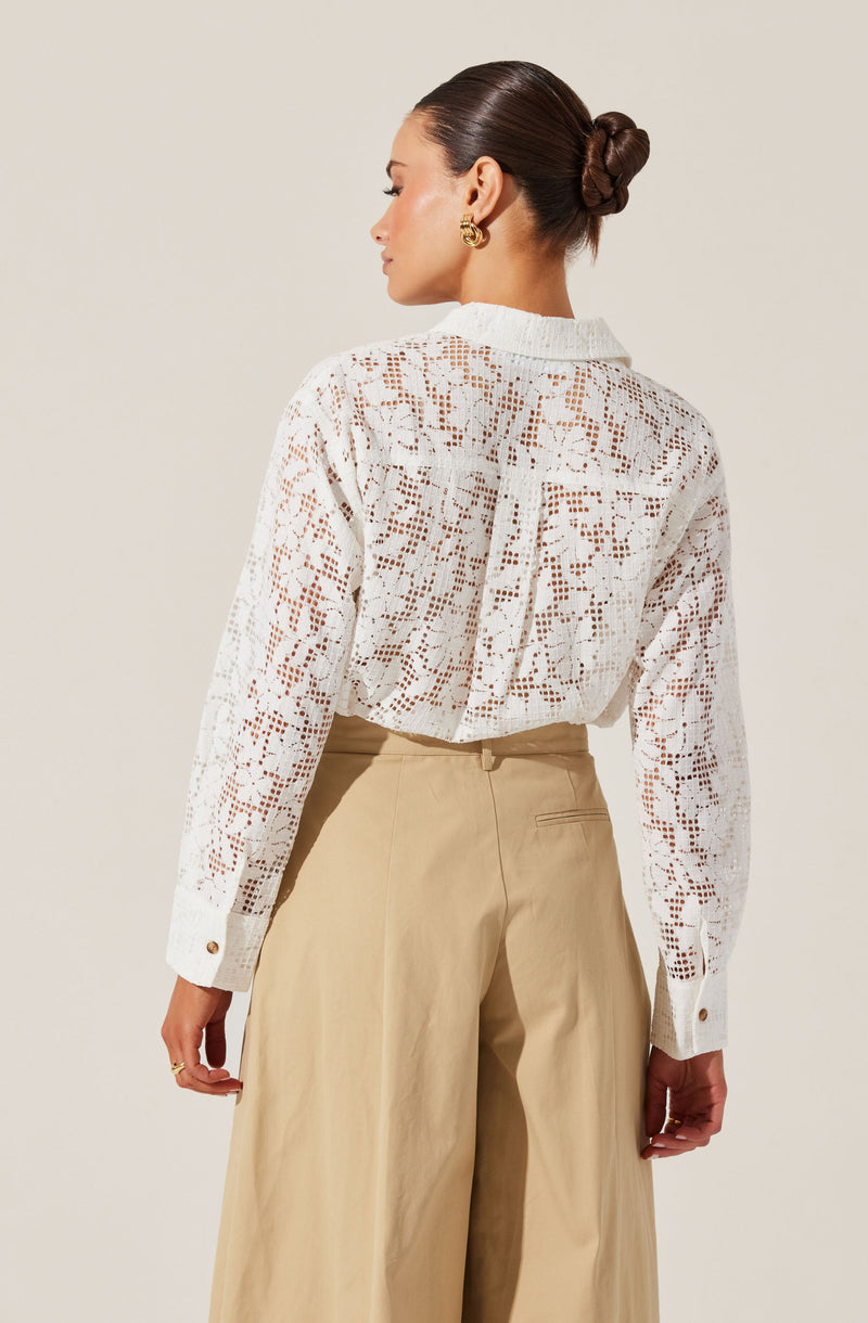 A woman is wearing a white, long-sleeved, openwork lace blouse with a high collar and buttoned cuffs, paired with high-waisted, wide-leg beige trousers. Her hair is styled in a sleek, low bun, and she has gold hoop earrings.