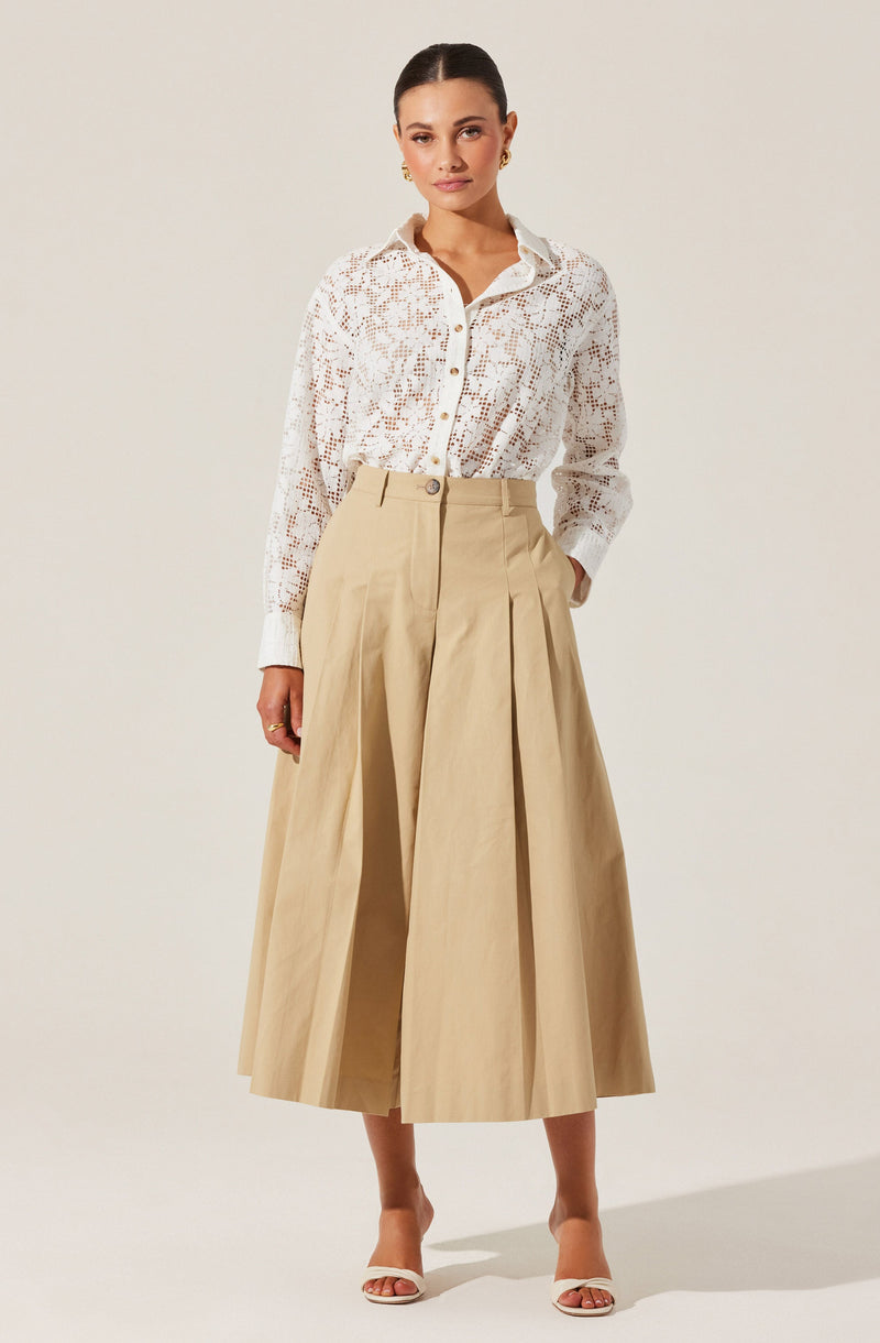 A woman is wearing a white, long-sleeved, button-up shirt with a lace or crochet pattern, paired with high-waisted, wide-leg beige trousers that feature pleats. She is also wearing white open-toe heels and small earrings.
