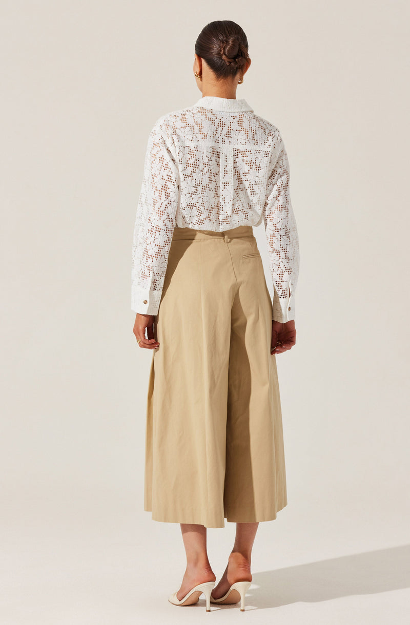 A woman is wearing a long-sleeved white blouse with a crochet or lace pattern, featuring a collar and buttoned cuffs. She pairs it with high-waisted, wide-leg beige trousers that reach mid-calf. She completes the outfit with white heeled shoes.