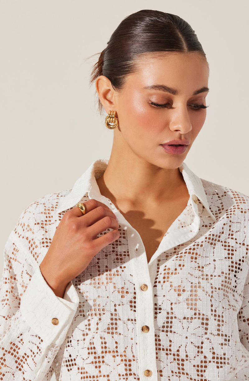 A woman is wearing a white, long-sleeved, button-up shirt with intricate lace or crochet cut-out patterns throughout the fabric. The shirt has a collar and small, round, light-colored buttons down the front.