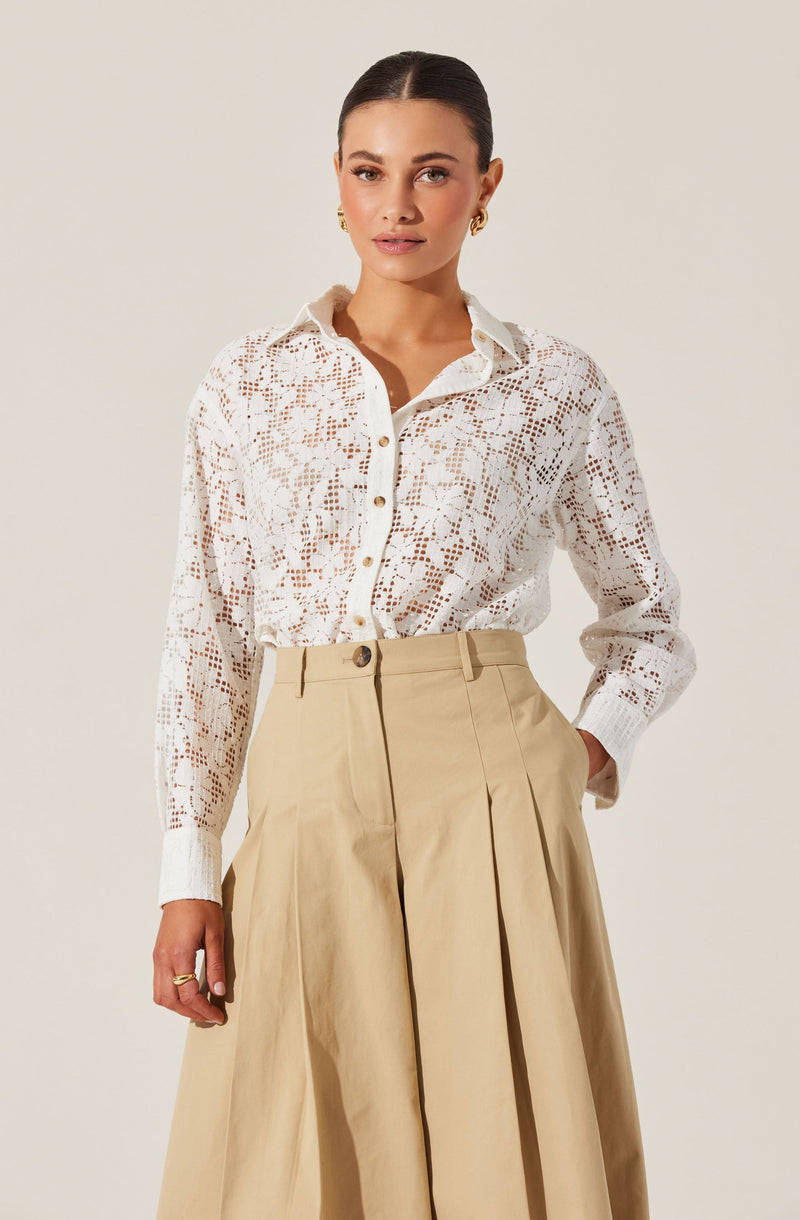 A woman is wearing a long-sleeved, white lace button-up shirt with a floral pattern and a classic collar. She has paired it with high-waisted, wide-leg beige trousers that feature pleats and a belt loop.