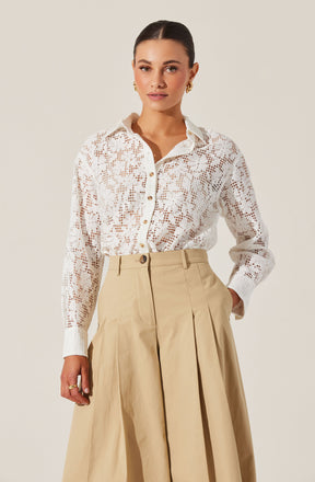 A woman is wearing a long-sleeved, white lace button-up shirt with a floral pattern and a classic collar. She has paired it with high-waisted, wide-leg beige trousers that feature pleats and a belt loop.