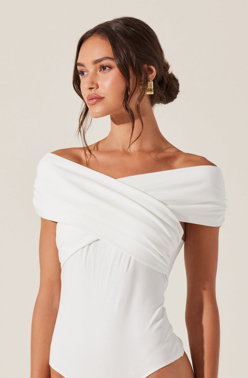 A woman is wearing an elegant white off-the-shoulder dress with wide, pleated fabric draped across the shoulders and bodice, creating a sophisticated look. She accessorizes with gold statement earrings and has her dark hair styled in a low, loose bun with soft waves framing her face.