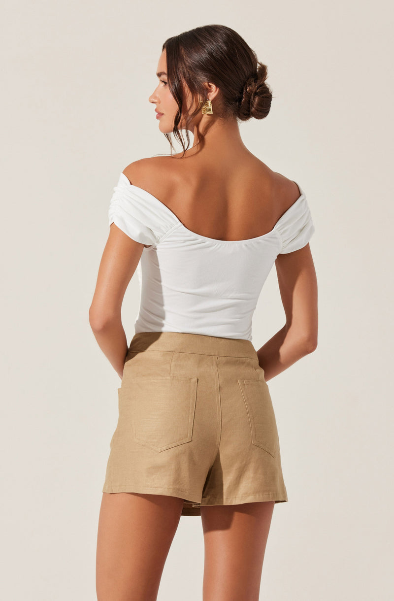 A woman is wearing an off-the-shoulder white top with short, ruched sleeves and a gathered neckline, paired with high-waisted beige shorts that have visible back pockets and a slightly loose fit.