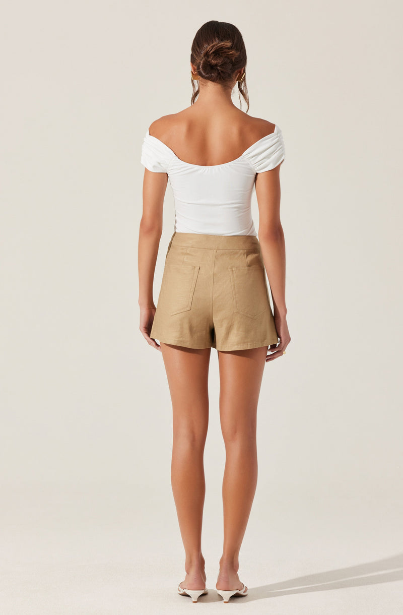 A woman is wearing a white off-the-shoulder top with ruched sleeves and a fitted bodice, paired with high-waisted beige shorts that have large back pockets. She is also wearing beige open-toe shoes. Her hair is styled in a loose updo.