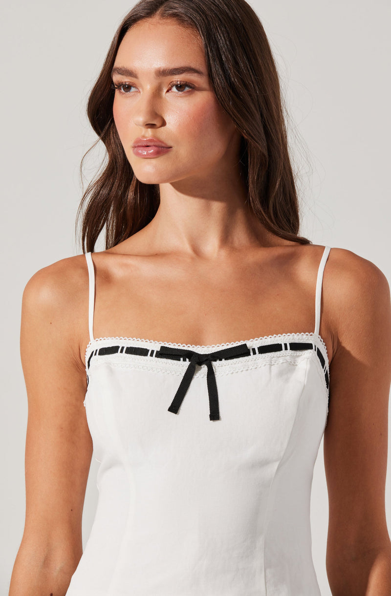 A woman is wearing a white sleeveless top with thin shoulder straps. The top features black ribbon details along the neckline with a small black bow in the center. The fabric appears lightweight and comfortable.