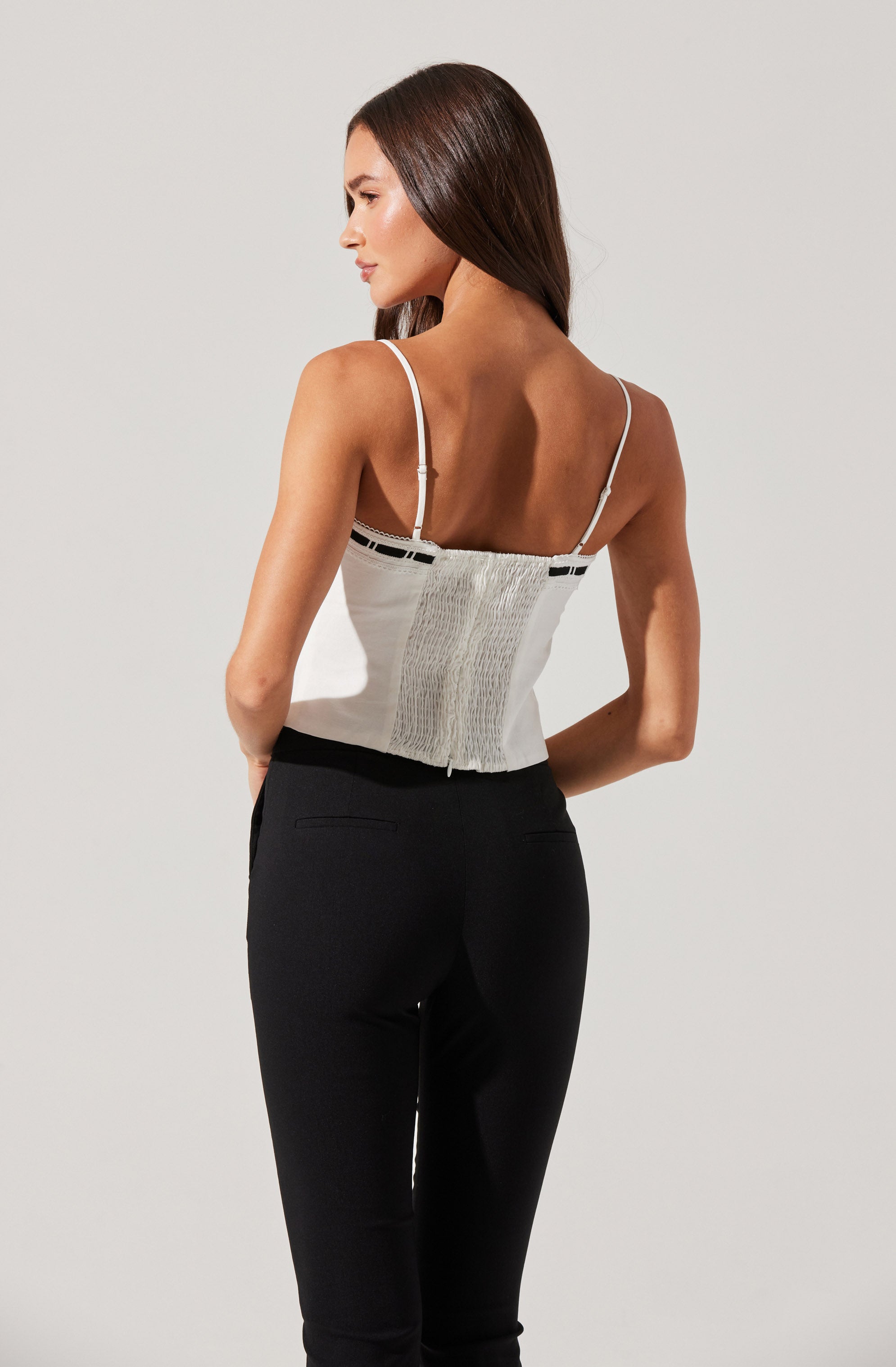 A woman is wearing a sleeveless, fitted top with thin spaghetti straps. The top is primarily white with black accents and features a textured, smocked panel in the center. She pairs it with high-waisted, black pants that have a slim fit.