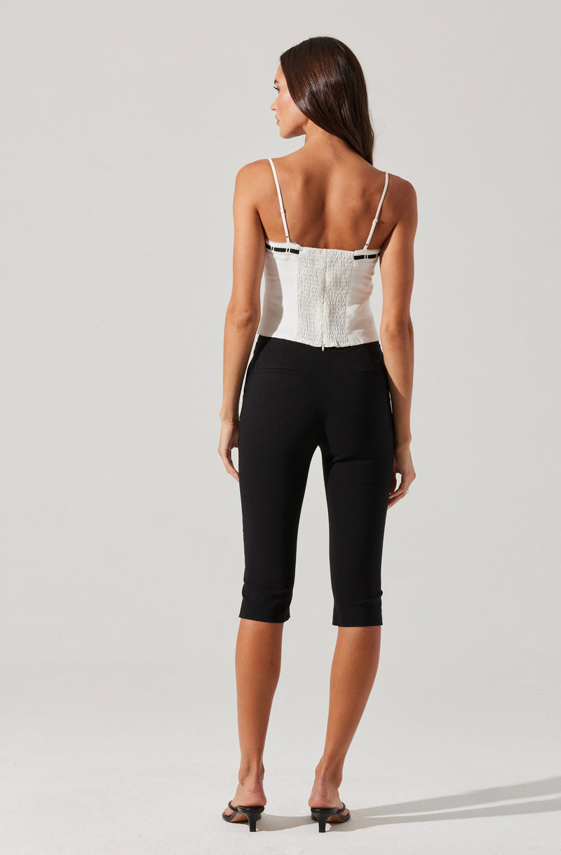 A woman is wearing a white sleeveless top with thin shoulder straps, featuring a fitted bodice with textured vertical panels and black accents near the chest area. She pairs it with black cropped pants that fit snugly and end below the knees, with a slight cuffed hem. She is also wearing black open-toe high heel sandals.