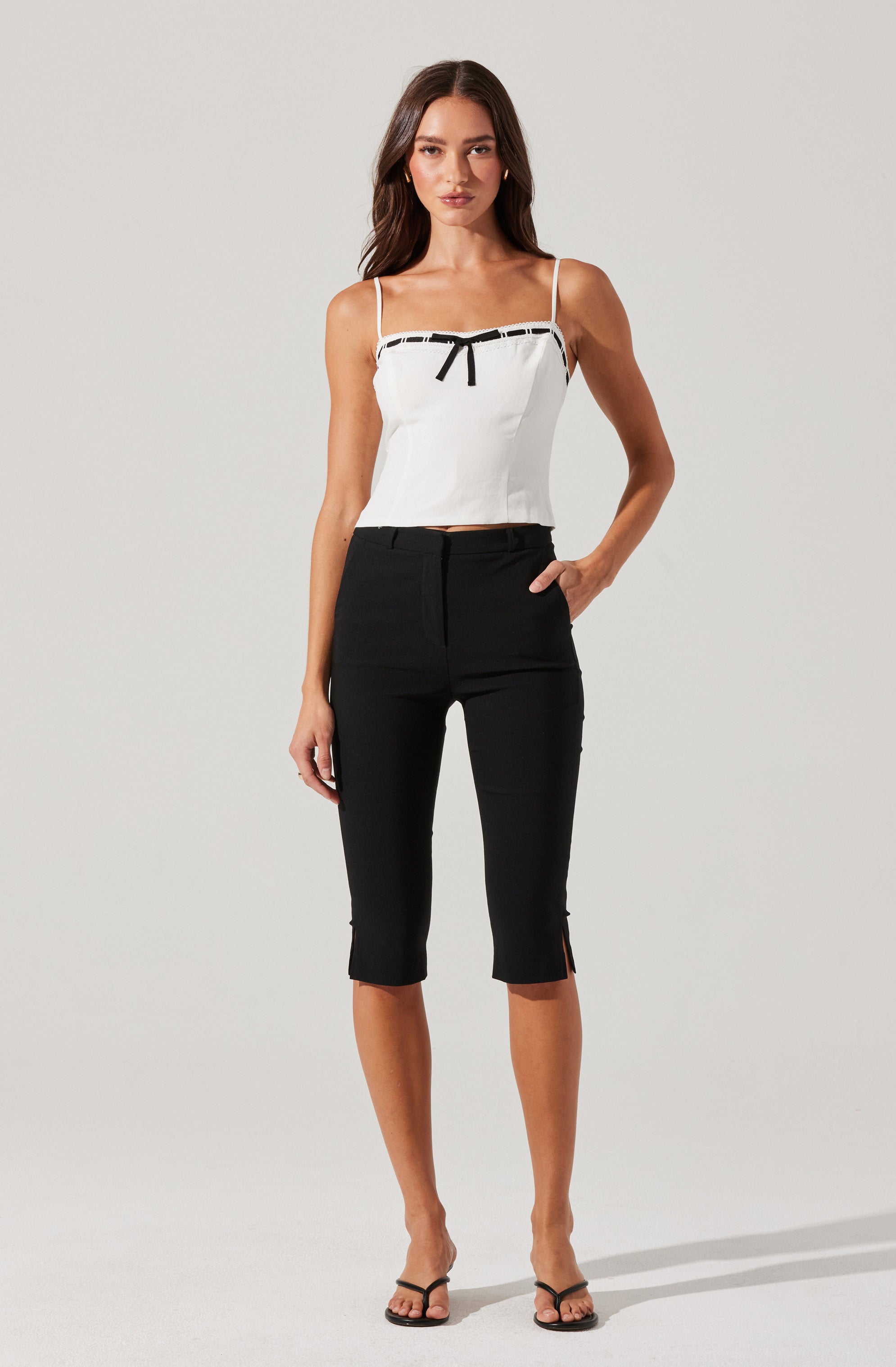 A woman is wearing a white sleeveless top with thin spaghetti straps, a black ribbon bow at the center, and black trim along the top edge. She is paired with black capri pants that have a slim fit, a high waist, and small side slits at the hem.