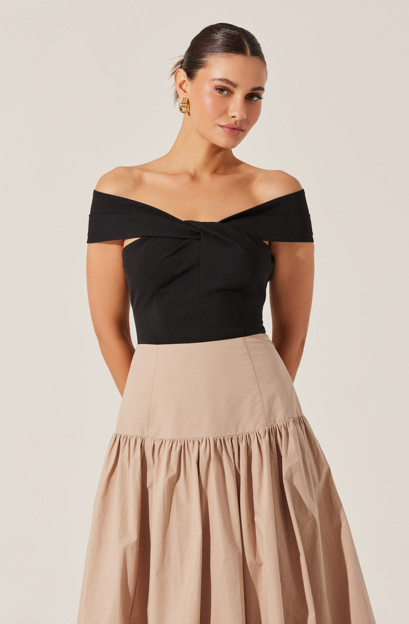 A woman is wearing an off-the-shoulder black top with a twisted knot detail at the front. She has paired it with a beige, high-waisted, A-line skirt that features a gathered ruffle at the hem. She accessorizes with gold hoop earrings.