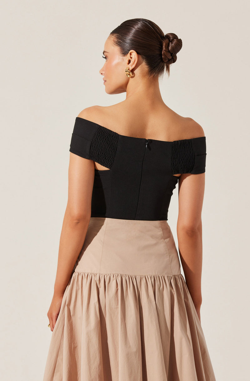 A woman is wearing an off-the-shoulder black top with textured, ruched detailing on the sleeves and a back zipper closure. She pairs it with a high-waisted, beige skirt that has a fitted waist and a full, gathered lower section. She accessorizes with gold hoop earrings.