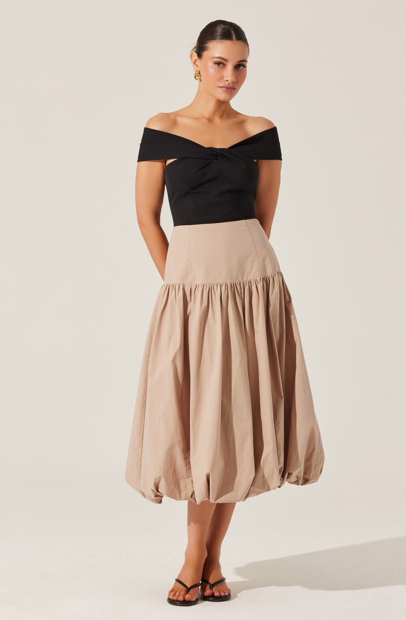 A woman is wearing an off-the-shoulder black top with a twist detail in the front. She pairs it with a high-waisted, beige, balloon-style skirt that is gathered at the waist and extends to mid-calf, creating a voluminous silhouette. She is also wearing black strappy sandals.