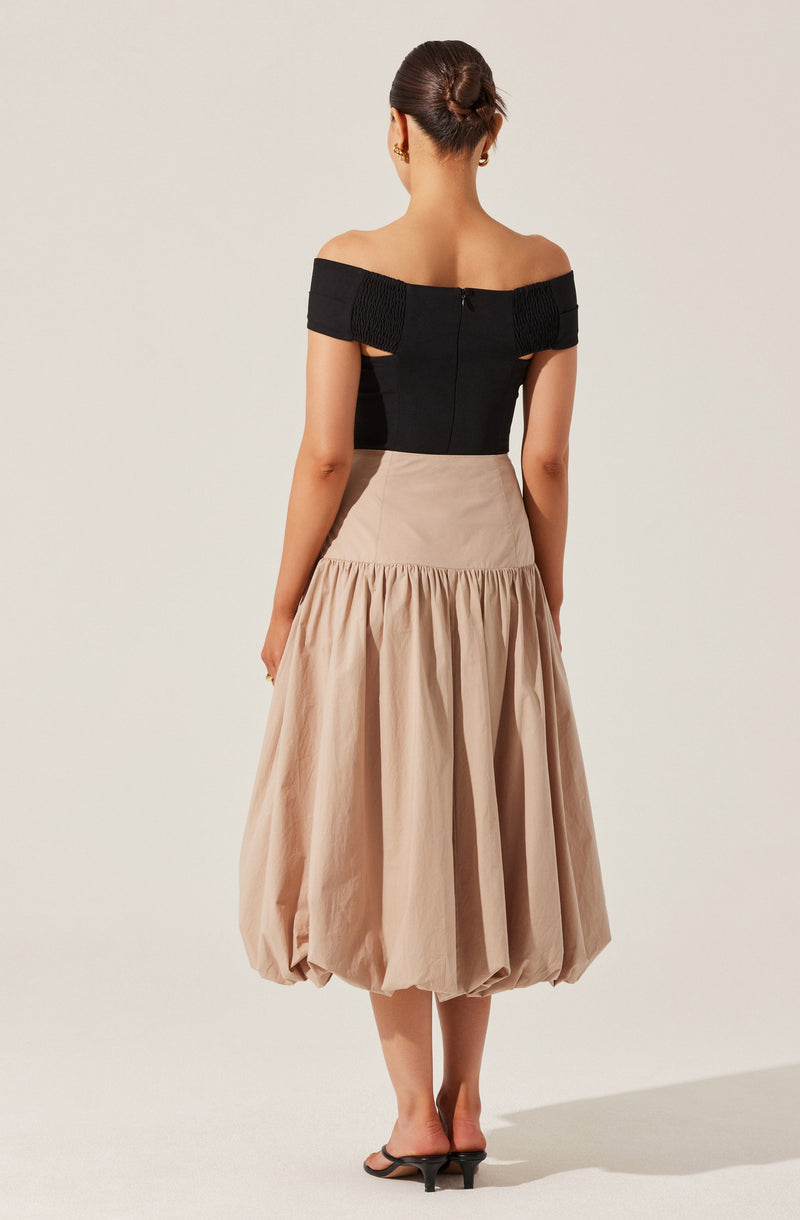 The woman is wearing an off-the-shoulder black top with textured band details on the sleeves, paired with a high-waisted, beige, gathered midi skirt that has a voluminous, bubble-like hem. She is also wearing black heeled sandals.