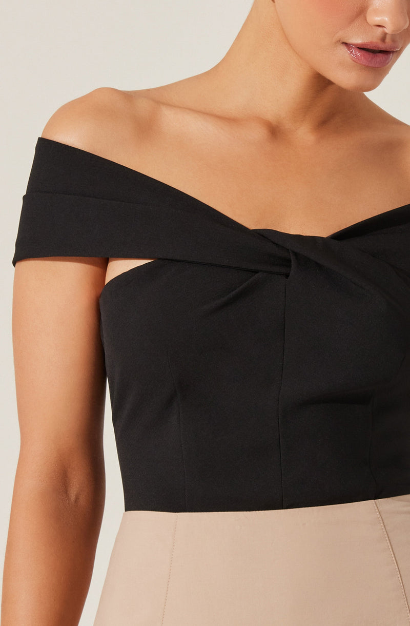 A woman is wearing an off-the-shoulder black top with wide, folded-over sleeves that drape over her upper arms. The dress features a twisted knot detail at the neckline, creating a gathered effect. The top is fitted and sleeveless on the shoulders, highlighting her collarbones and upper arms. She is also wearing a light-colored skirt.