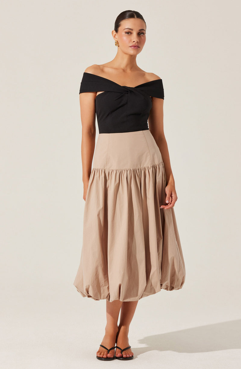A woman is wearing an off-the-shoulder black top with a twisted knot detail at the center of the neckline. She pairs this with a high-waisted, beige, voluminous skirt that has a gathered, bubble hem ending just above her ankles. She completes her look with gold earrings and black strappy sandals.