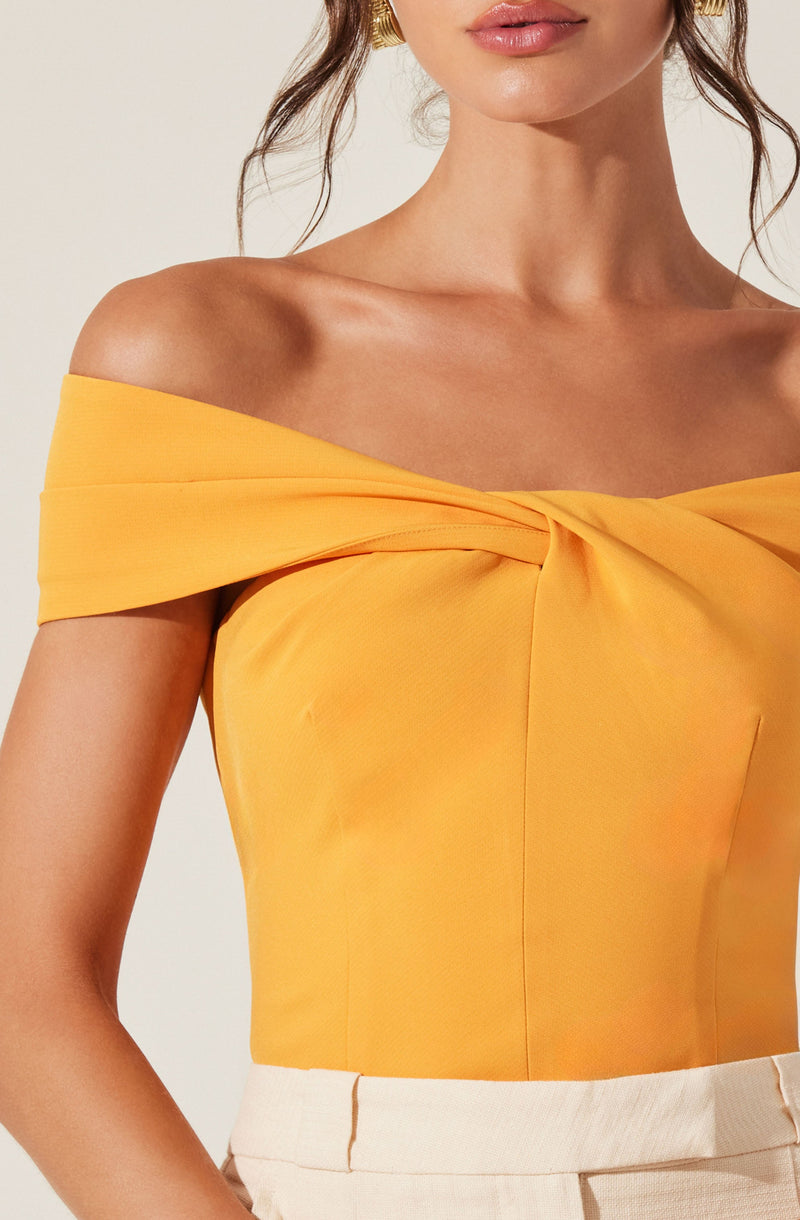 A woman is wearing an off-the-shoulder, bright yellow top with a twisted knot detail at the neckline. The top has short, wide sleeves that drape over her shoulders. She pairs the top with high-waisted, light beige pants.