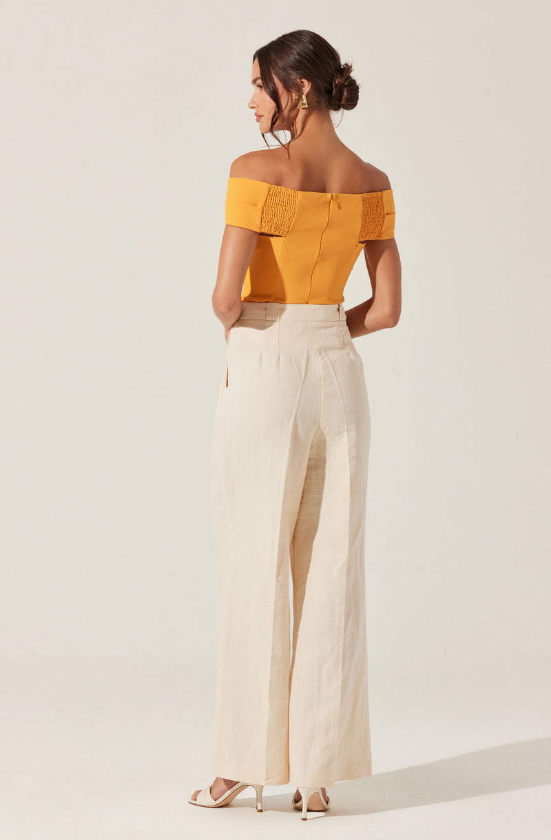 A woman is wearing a bright yellow off-the-shoulder top with gathered detail at the sleeves and a fitted back. She pairs it with high-waisted, wide-leg cream-colored trousers made from a light, textured fabric. She is also wearing nude high-heeled sandals.