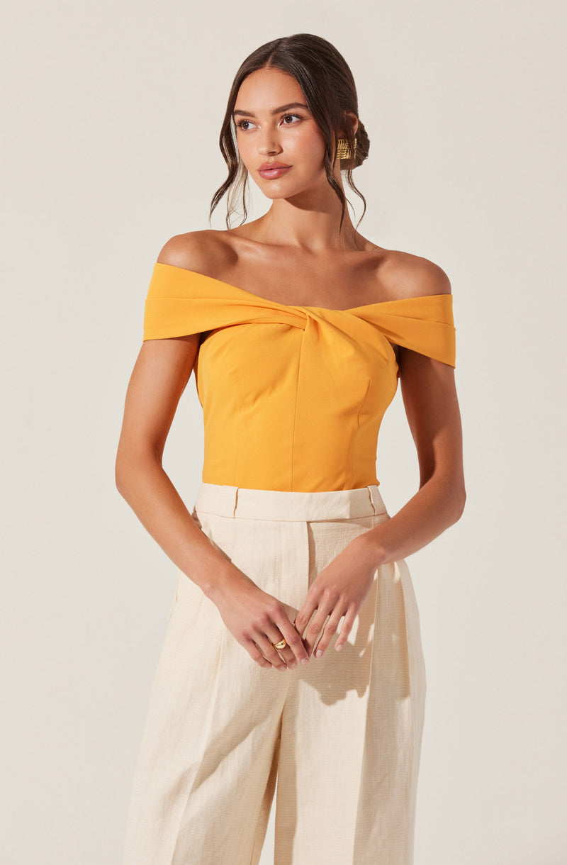 A woman is wearing an off-the-shoulder yellow top with a twisted knot detail at the front. She pairs it with high-waisted, wide-leg cream trousers. She has long, dark hair styled in loose waves and is accessorized with gold earrings.