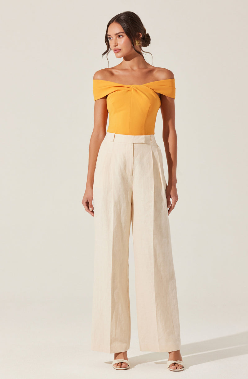 A woman is wearing an off-the-shoulder, bright yellow top with a twisted knot detail at the front, paired with high-waisted, wide-leg cream-colored trousers. She has on open-toe heels and gold earrings.