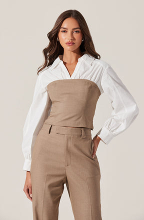 A woman wearing a white shirt with long, puffed sleeves and buttoned cuffs, layered with a beige strapless bustier top over the shirt. She is also dressed in matching beige high-waisted trousers with belt loops and a front zipper.