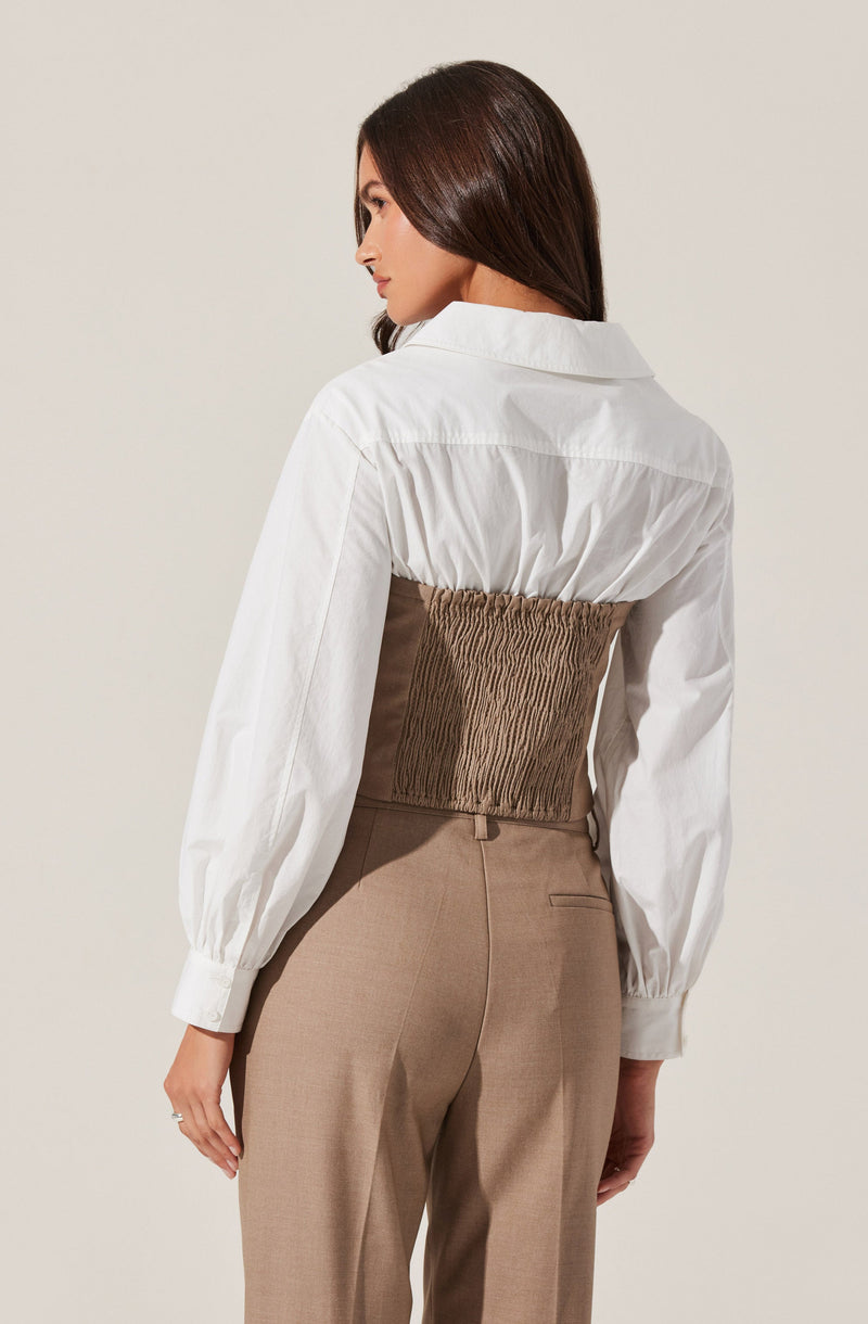 A woman is wearing a white long-sleeve button-up shirt with a wide collar and puffed sleeves. She has a brown, elasticized, ruched waist corset over the shirt. She is also wearing high-waisted beige trousers.