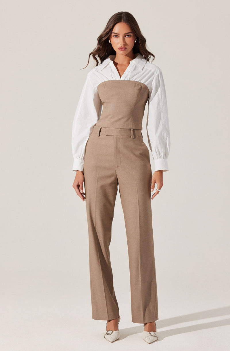 A woman is wearing a white, long-sleeved, collared shirt with vertical stripes. Over the shirt, she has on a strapless, beige tailored jumpsuit that fits snugly at the top and flows into wide-leg trousers. She is also wearing pointed white heels.