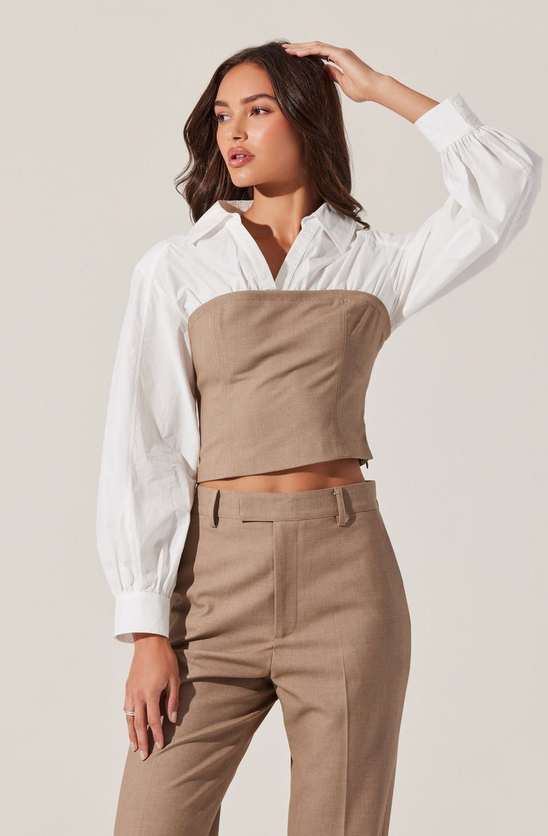 A woman is wearing a white long-sleeve button-up shirt with puffed sleeves and wide cuffs. Over the shirt, she has on a tan, strapless, cropped bustier-style top. She is also dressed in high-waisted tan trousers with belt loops.