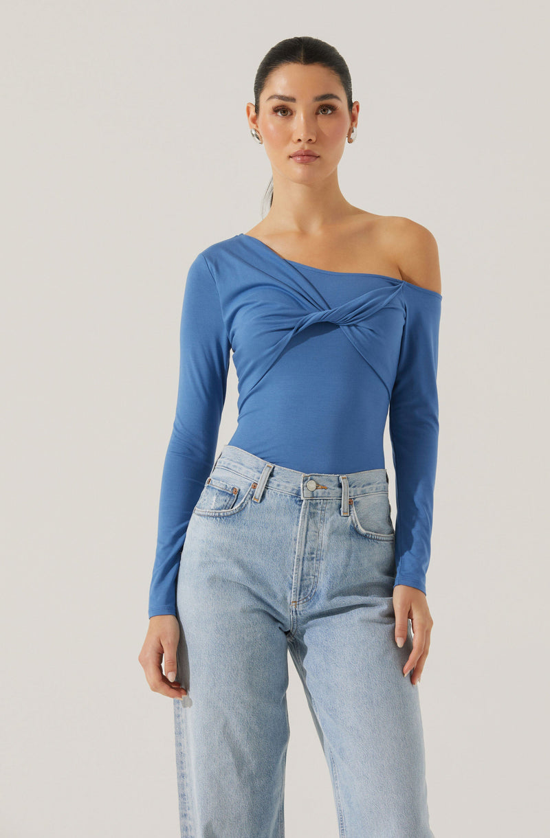 A woman is wearing a blue, off-the-shoulder long-sleeve top with a twisted knot detail on the front, paired with high-waisted light-wash jeans.