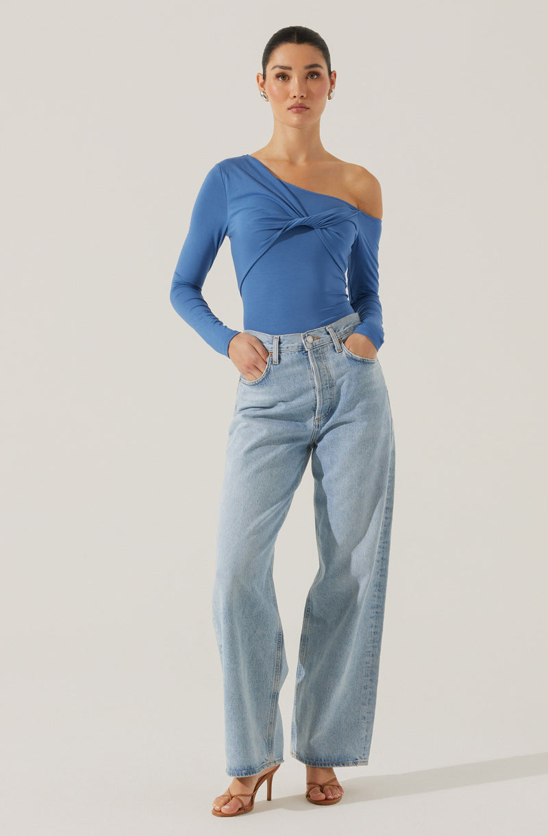 A woman is wearing a blue, asymmetrical, long-sleeved top with a twisted knot detail across the chest and an off-the-shoulder neckline on the right side. She pairs the top with high-waisted, light-wash, wide-leg jeans. She is also wearing nude-colored high heels.