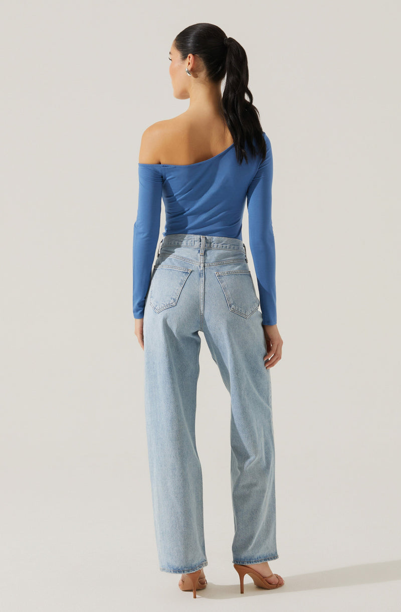A woman is wearing a fitted, long-sleeve, off-the-shoulder blue top with a ruched detail on the side. She pairs it with high-waisted, light-wash, straight-leg jeans that have a classic five-pocket design and slightly frayed hems. She completes the outfit with nude high-heeled sandals.